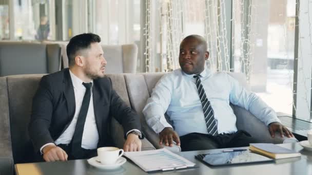 Two multi-ethnic businessmen discussing startup ideas in glassy cafe during lunch time. Coffee cup, financial graphs and digital tablet are on table — Stock Video