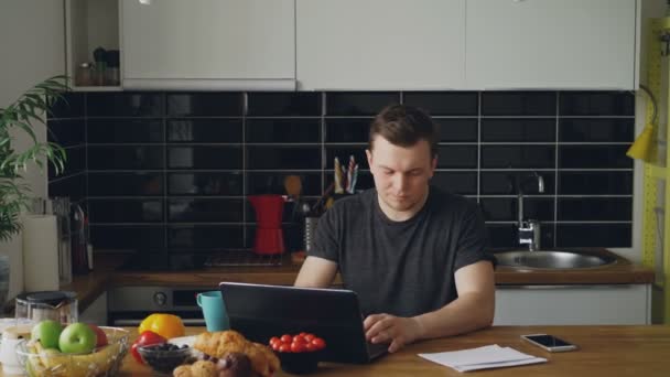 Bonito jovem receba boas notícias lendo carta de papel na cozinha enquanto toma café da manhã em casa — Vídeo de Stock