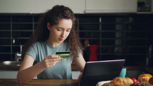 Joyeux jeune femme ayant des achats en ligne à l'aide d'un ordinateur portable et carte de crédit pendant le petit déjeuner dans la cuisine à la maison — Video