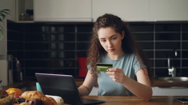 Весела молода жінка, яка купує онлайн за допомогою ноутбука та кредитної картки, снідаючи на кухні вдома — стокове відео