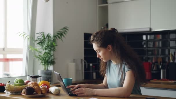 Jovem mulher alegre concentrada trabalhando na cozinha digitando computador portátil durante o café da manhã — Vídeo de Stock