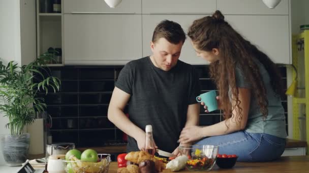 Happy νεαρό ζευγάρι μαγείρεμα και κουβεντιάζοντας, ενώ ευτυχώς άνθρωπος κοπής λαχανικών για πρωινό στην κουζίνα στο σπίτι. Σχέσεις και οικογένεια έννοια — Αρχείο Βίντεο