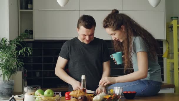 Glada unga par matlagning ant prata i köket hemma. Attraktiv man utfodra sin flickvän medan skära grönsaker till frukost på morgonen — Stockvideo