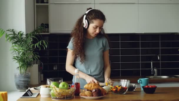 Привлекательная счастливая девушка в наушниках танцы и и слушать музыку во время приготовления здорового овощного салата на кухне дома по утрам — стоковое видео