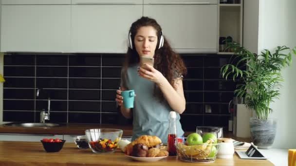 Привлекательная счастливая девушка танцует на кухне, просматривая социальные сети на смартфоне и слушая музыку дома утром — стоковое видео