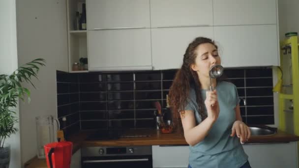 Vrolijke jonge grappige vrouw dansen en zingen met pollepel terwijl het hebben van vrijetijd in de keuken thuis — Stockvideo