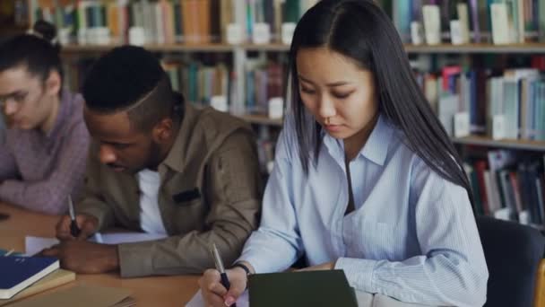 Concentrada asiática estudiante preparándose para el examen y escribir notas mientras está sentada en la mesa en la biblioteca universitaria con sus compañeros de clase estudiando libros — Vídeos de Stock