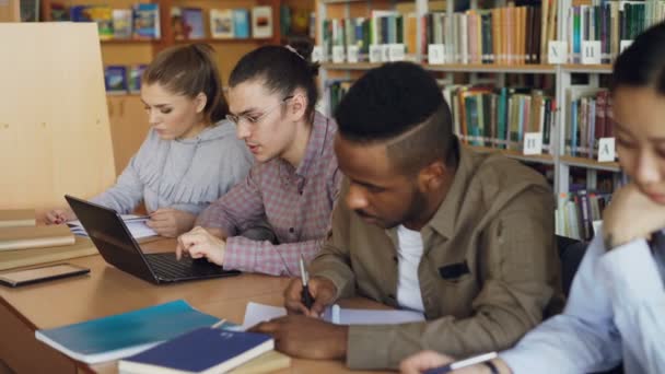 Concentre-se em estudante hipster masculino em óculos usando o computador portátil enquanto se prepara para exames na biblioteca da universidade com seus colegas de classe — Vídeo de Stock