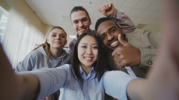 Ein asiatisches Mädchen hält ein Smartphone in der Hand, macht Selfie-Fotos mit fröhlichen multiethnischen Klassenkameraden und hat Spaß in der Universitätsbibliothek. Frau zeigt Berührungsängste und sieht Bilder — Stockvideo