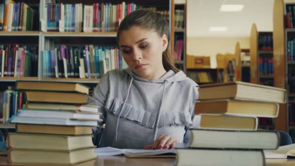 Концентрированная молодая студентка читает книги и пишет в блокноте во время работы в школьном проекте в университетской библиотеке — стоковое видео