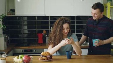 genç çekici kıvırcık mutlu beyaz kadın mutfak manifatura smartphone yoluyla oturuyor, kocası geliyor ve o telefonu komik bir şey gösterir