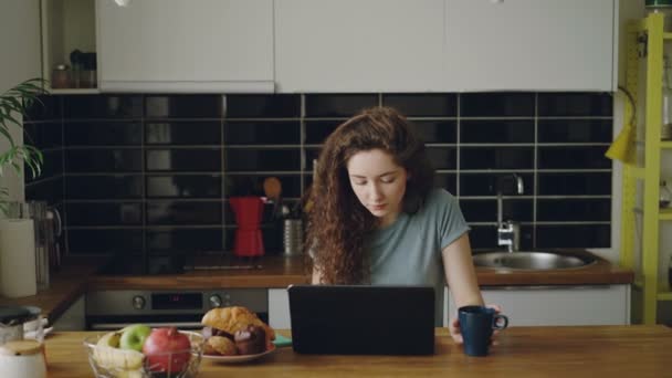 Jeune femme caucasienne bouclée réfléchie est assise dans la cuisine à l'intérieur devant un ordinateur portable, buvant du café et travaillant, elle jette un coup d'oeil dans la fenêtre — Video
