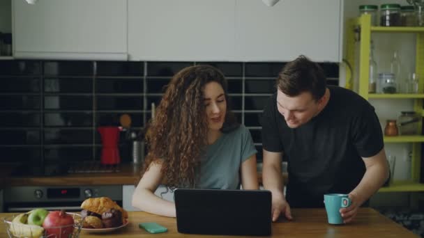 Jeune femme bouclée caucasienne positive assis ar table dans la cuisine travaillant sur un ordinateur portable, son mari vient et elle lui montre quelque chose dans un ordinateur portable, ils sourient et en discutent — Video