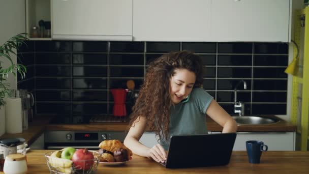 Jeune femme caucasienne bouclée assez positive qui travaille sur un ordinateur portable et parle au téléphone assis dans la cuisine moderne, elle imprime et parle en même temps — Video