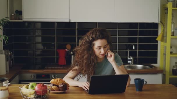 Jeune femme caucasienne assez sérieuse bouclée qui travaille sur un ordinateur portable et parle au téléphone assis dans la cuisine moderne, elle imprime et parle en même temps — Video