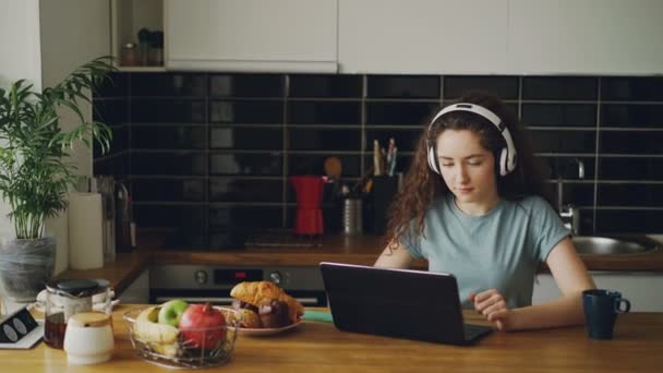 积极快乐的白种人漂亮卷曲十几岁的女孩坐在桌子上聊天与某人在笔记本电脑上听音乐在厨房里的臭虫耳机 — 图库视频影像
