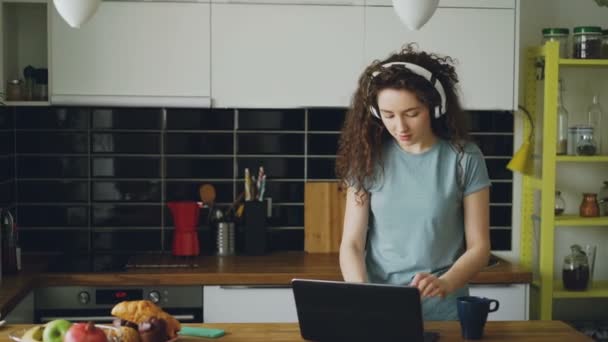 Jovem adolescente usando fones de ouvido aparece na cozinha chegando à mesa, bebendo chá da xícara, dançando e imprimindo algo no laptop — Vídeo de Stock