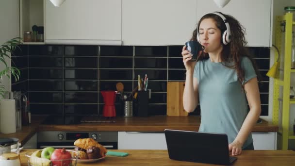 Jeune belle belle femme bouclée caucasienne portant des écouteurs dansant dans la cuisine près de la table devant l'ordinateur portable, imprimer quelque chose, prendre le téléphone et envoyer des SMS — Video