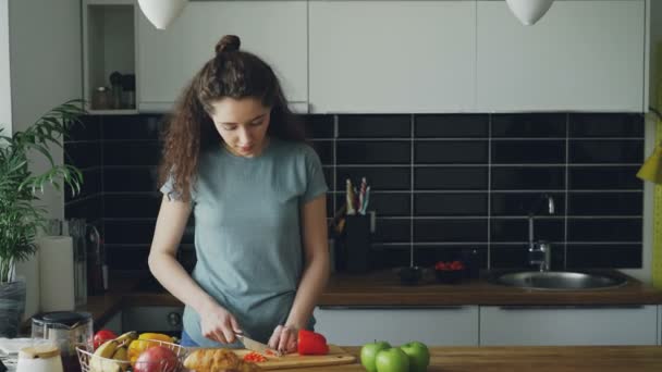Lockigt kvinna dans nära tabell skärande röd paprika i köket hemma, hennes make kommer anläggningen cup, visar henne något roligt i smartphone och de ler — Stockvideo