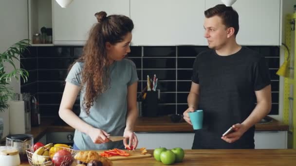 Jonge Kaukasische paar luisteren naar muziek op smartphone, dansen in de buurt van tafel in keuken thuis, mooie vrouw is het kappen van rode peper voor salade, man met cup — Stockvideo
