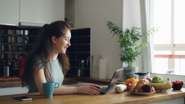 Curly donna caucasica abbastanza positivo seduto a tavola skyping sul computer portatile, sta parlando seduto in cucina vicino alla finestra, tazza di caffè in piedi vicino al computer portatile — Video Stock