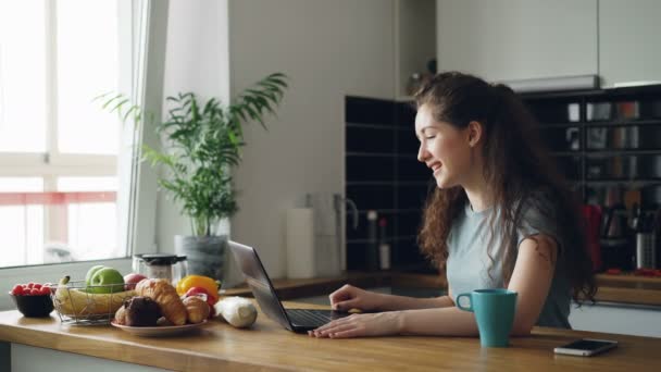 Kudrnatá docela pozitivní Kavkazský žena sedí u stolu skyping na notebooku, se mluví, seděl v kuchyni u okna, šálek kávy stojící poblíž notebooku — Stock video