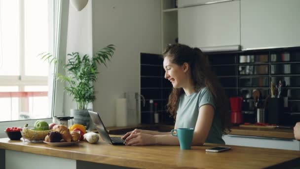 Mladí kudrnaté docela pozitivní Kavkazský žena sedí u stolu a mít video hovor na přenosném počítači, ona sedí v kuchyni v blízkosti okna, její přítel přijde a že skype společně v pozitivním způsobem — Stock video