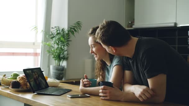 Young beautiful caucasian couple sitting in modern kitchen at table in front of laptop skyping with two girls, they are smiling and laughing and waving hands — Stock Video