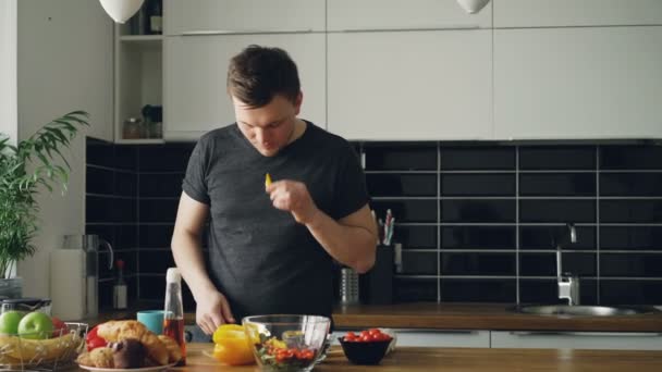 Ung snygg kaukasiska man står vid bordet skär grönsaker och försöker lappa av gul paprika, hans flickvän kommer rasande holding mobiltelefon, man skrattar — Stockvideo