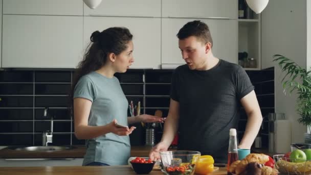 Młoda kobieta kręcone, pokazując coś nieprzyjemnego w mężów telefon podczas gdy on gotuje, one są krzyki i kłótnie, człowiek jest zły i podrażnionej — Wideo stockowe