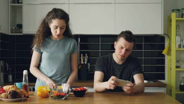 Привлекательная пара на кухне. Мужчина играет в видеоигры на смартфоне, пока его девушка готовит — стоковое видео