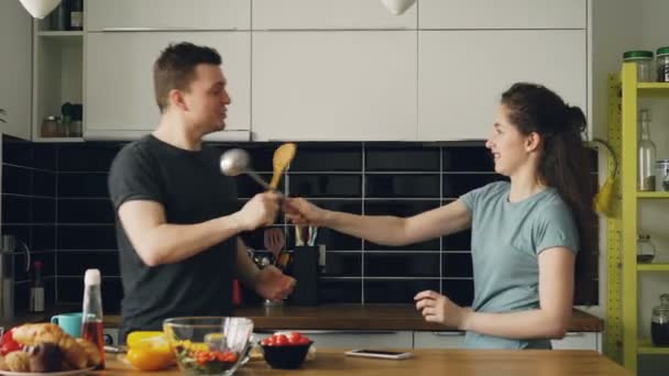 Pareja feliz divirtiéndose en la esgrima de la cocina con cucharas grandes mientras cocina el desayuno en casa — Vídeos de Stock