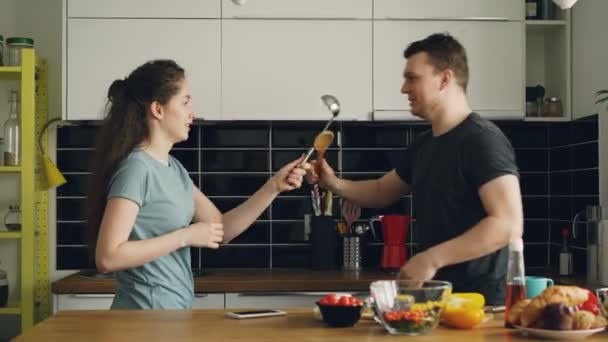 Счастливая пара веселится на кухне фехтование с большими ложками во время приготовления завтрака дома — стоковое видео