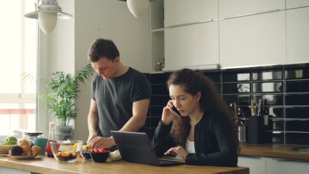 Σοβαρή Καυκάσιος ωραίο ζευγάρι στην κουζίνα, ο άνθρωπος κοπή σαλάτα, στέκεται σιωπηλή γυναίκα κάθεται στο τραπέζι εργασίας για φορητό υπολογιστή και να μιλάμε στο τηλέφωνο — Αρχείο Βίντεο