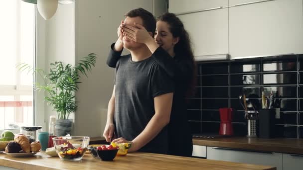 Femme d'affaires caucasienne bouclée debout derrière son mari dans la cuisine le faisant deviner, il ouvre les yeux, ils rient et sourient discuter de sa cuisine — Video