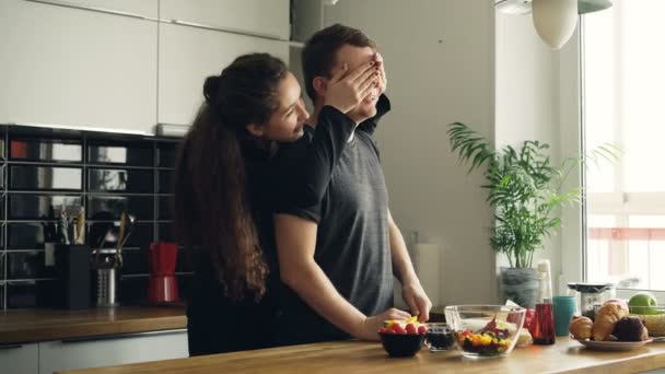 Curly mulher de negócios caucasiana de pé atrás de seu marido na cozinha fazendo-o adivinhar, ele abre os olhos, eles estão rindo e sorrindo discutindo sua culinária — Vídeo de Stock