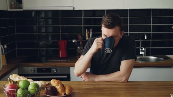 Портрет молодого кавказького чоловіка, що сидить за столом у сучасній просторій кухні, дивиться через вікно, п'є чай, втомився і засмучений, закриваючи очі руками — стокове відео