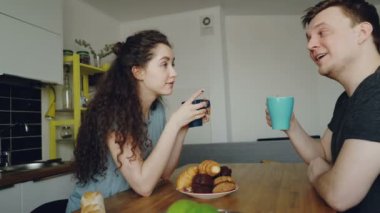 bir şey onun evde modern mutfakta masada oturan genç çift, kıvırcık kadın çay içme kamera oturuyor ve croussant yemek, bardak tutuyorlar