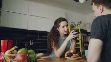 bir şey onun evde modern mutfakta masada oturan genç çift, kıvırcık kadın çay içme kamera oturuyor ve croussant yemek, bardak tutuyorlar