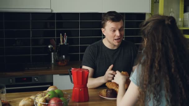 Jonge Kaukasische paar zittend aan tafel in de moderne keuken thuis iets dicussing, krullend vrouw zit terug naar camera thee drinken en eten croussant, houden ze cups — Stockvideo