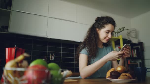 Jovem bonita encaracolado bonita caucasiana mulher sentada à mesa em boa cozinha usando smartphone, ela está enviando mensagens de texto para alguém e sorrindo, calma e feliz — Vídeo de Stock