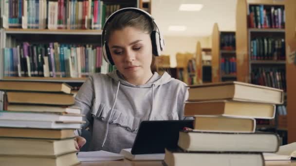 Une jeune femme souriante dans un casque blanc est assise à table dans une bibliothèque universitaire, tenant une tablette numérique et écoutant de la musique. Elle danse un peu et écrit des conférences dans un cahier — Video