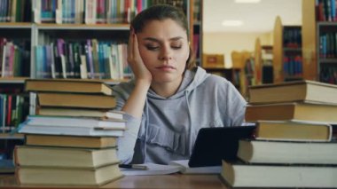 Güzel beyaz genç kadın Üniversite Kütüphanesi dijital tablet tutan masada oturuyor. Başını sallayarak ve defterini içinde yazma yorgun ve bitkin, kız