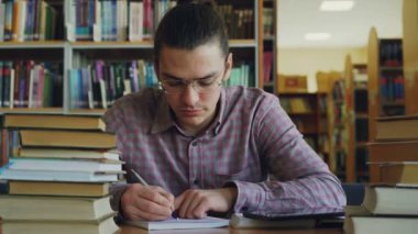 Defterini hesaplamalarda üzerinde Üniversitesi Kütüphane düşüncesinde masada oturan gözlüklü genç yakışıklı genç adam. Uzağa bakıyor ve witing thoughtfuly
