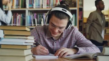 Genç yakışıklı beyaz öğrenci büyük okul Kütüphanesi masada oturan bir şey defterini içinde yazıyor. O gülümsüyor ve pozitif ve mutlu, masanın üzerinde yatan kitap görünüyor