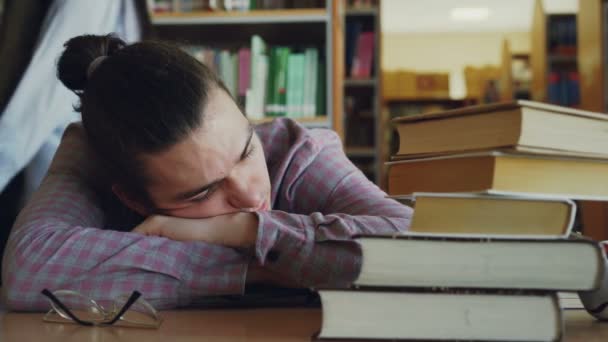 Guapo joven adolescente sentado con la cabeza sobre la mesa está durmiendo en la biblioteca de la escuela rodeado de enormes montones de libros. Sus gafas están tumbadas frente a él. — Vídeos de Stock