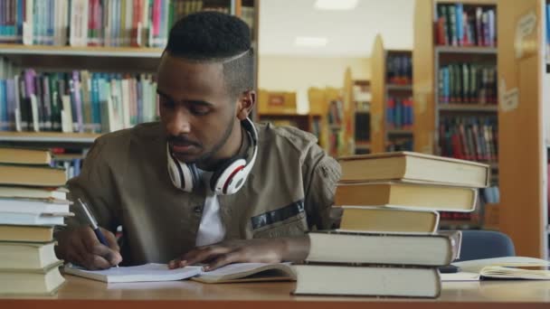 Positivo afroamericano joven guapo chico con grandes auriculares está sentado en la mesa con libros, mirando a través de la ventana, sonriendo y escribiendo en su copybook — Vídeos de Stock