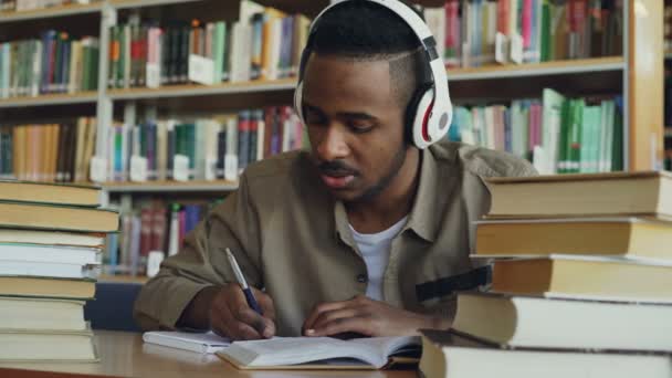 Guapo afroamericano estudiante masculino con auriculares está escuchando música sentada en la mesa en gran biblioteca espaciosa escritura conferencia rodeada de libros — Vídeo de stock