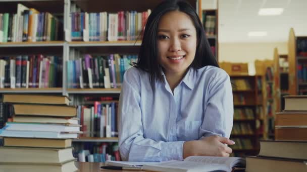 Portrét mladého krásná Asijská studentka sedí u stolu s piloty učebnic v knihovně při pohledu na fotoaparát. Směje se pozitivně. — Stock video