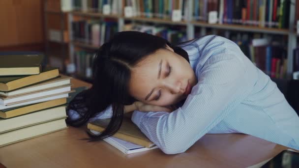 Молода красива втомлена азіатська студентка сидить за столом з головою на ньому, вона спить на підручнику, оточеному купами у великій університетській бібліотеці — стокове відео